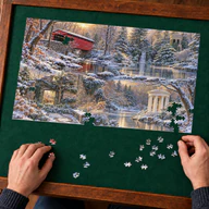 Lifestyle mockup of the 1,000-piece Gardens and Valleys of Greater Philadelphia Winter puzzle on a wooden puzzle table with hands beside loose pieces